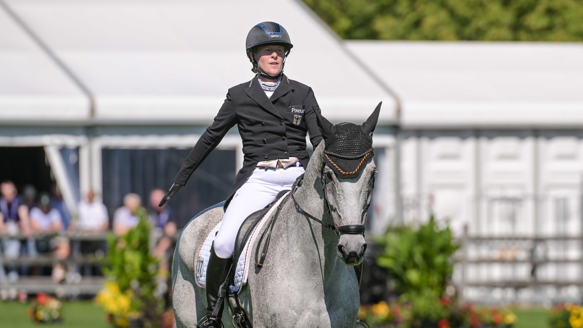 Image shows a rider in dressage clothing saluting the judges at the end of a dressage test. She is riding a grey horse, who wears an ear bonnet. There are white tents and spectators in the background