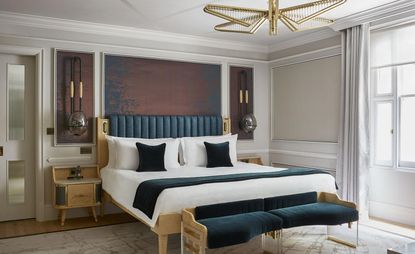 A room in the Mandarin Oriental Hyde Park. Walls with trimming are painted in light gray and beige. Bedframe and night tables are made from light wood, while the upholstery is in dark velvet blue. Golden light fixtures compliment the rest of the furniture. 