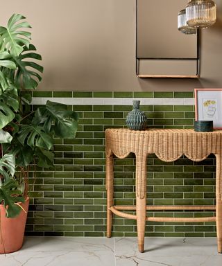 A bathroom with dark green subway tiles, a brown scalloped shelf with vases on it, a gold mirror on the wall, and a tall leafy green plant