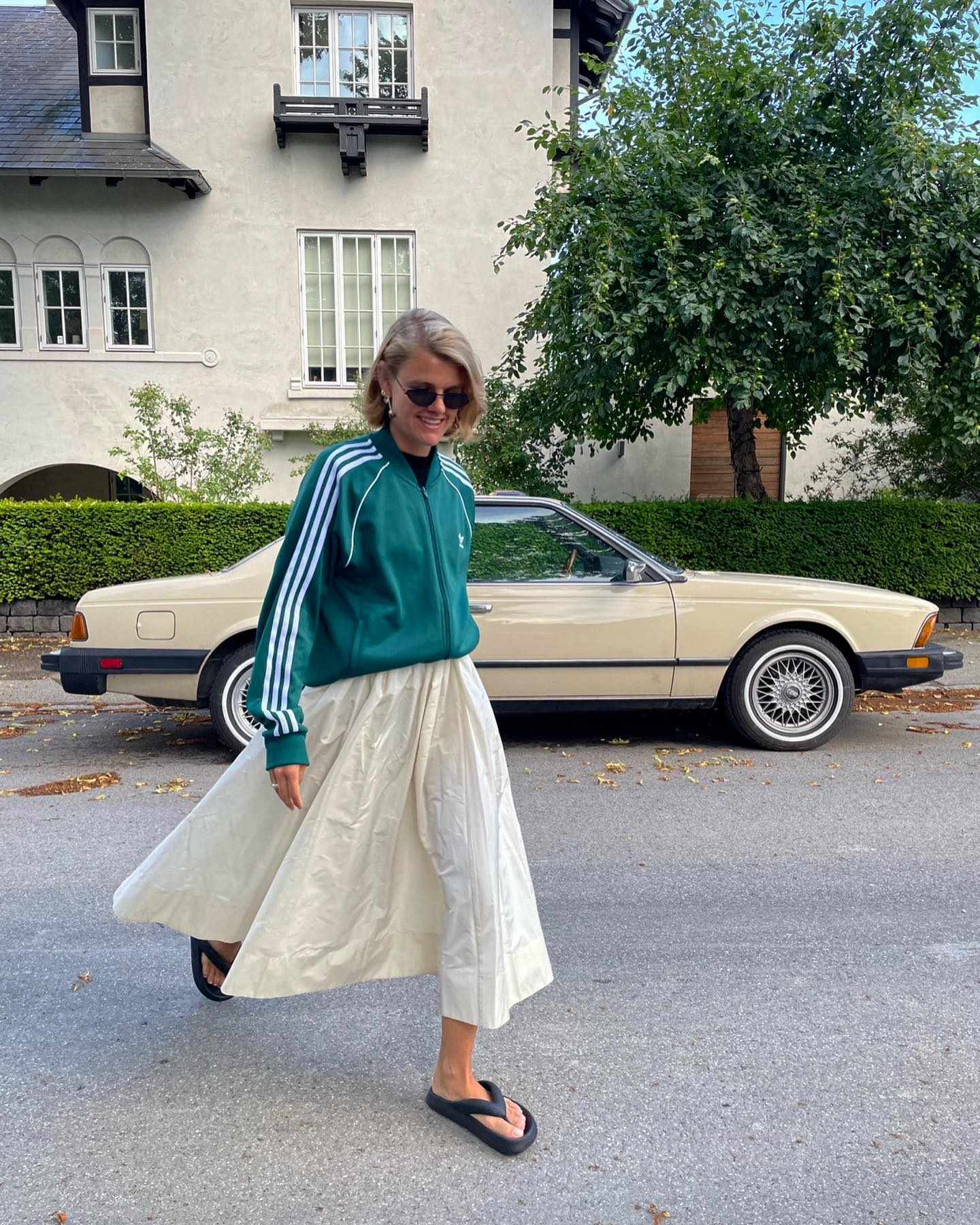 Scandi fashion influencer Amalie Neilsenn walks across a street in a sporty-chic outfit with a green Adidas zip-up jacket, full white skirt and platform flip-flop sandals