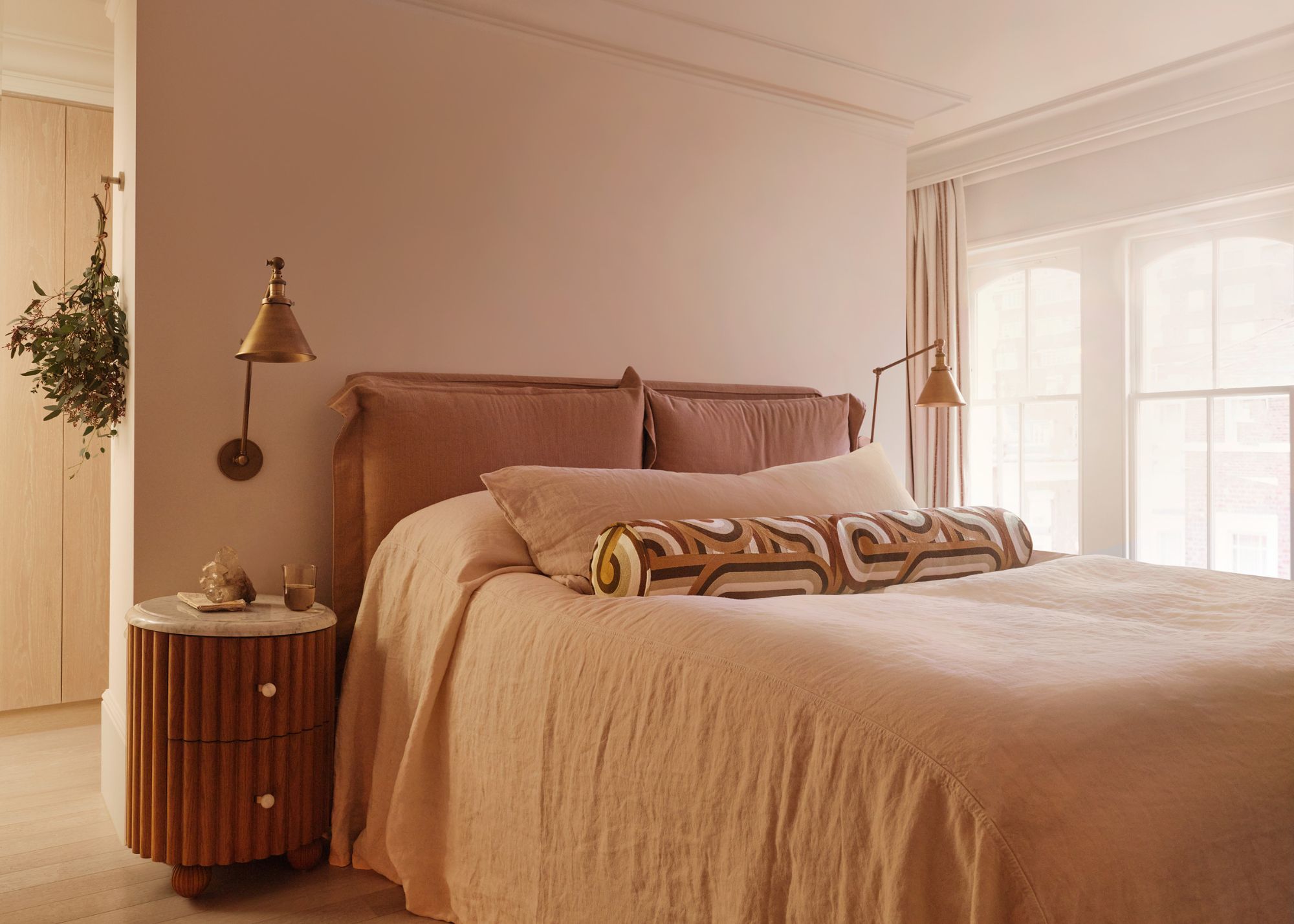 A neutral bedroom with soft pink walls, a large bed with natural linen bedding, a round nightstand, brass wall sconces, and a large window.