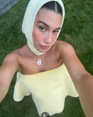 Hailey Bieber wearing a soft yellow headscarf, strapless Jacquemus dress, and B necklace.