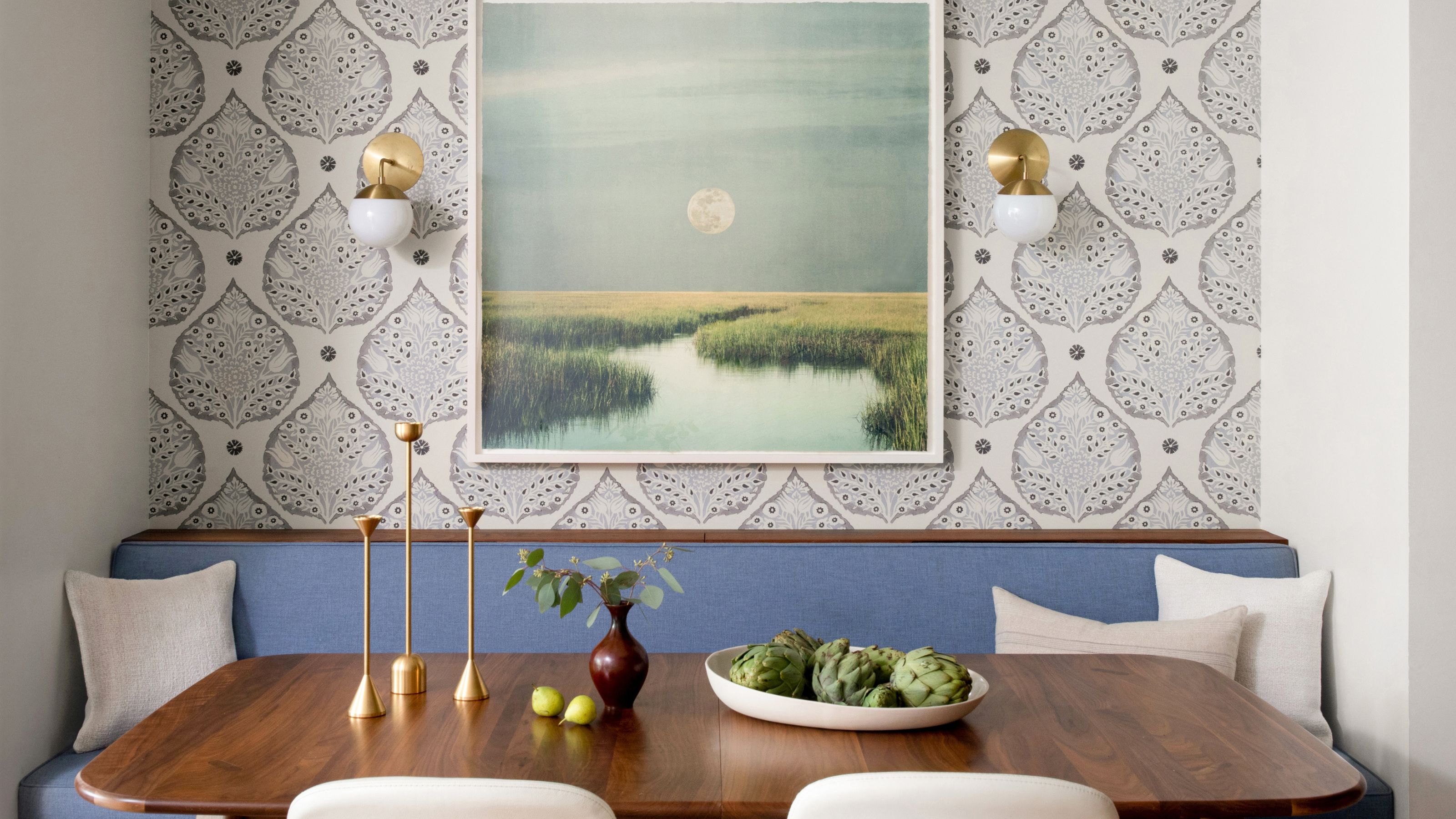 An apartment dining nook with neutral patterned wallpaper, powder blue banquette seating, a dark wood table, and light neutral cushions. A landscape artwork on the walls with two brass sconces. 