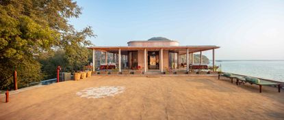 Daytime front entrance image of the RAAS Chhatrasagar, neutral pathway with two lake view seats to the right, trees to the left, lake to the right, hilly mountain to the back, blue sky