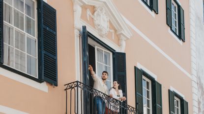 A man, who is pointing, and a woman, on the balcony of the salmon-pink hotel Can Alberti 1740, with green-shuttered windows on either side