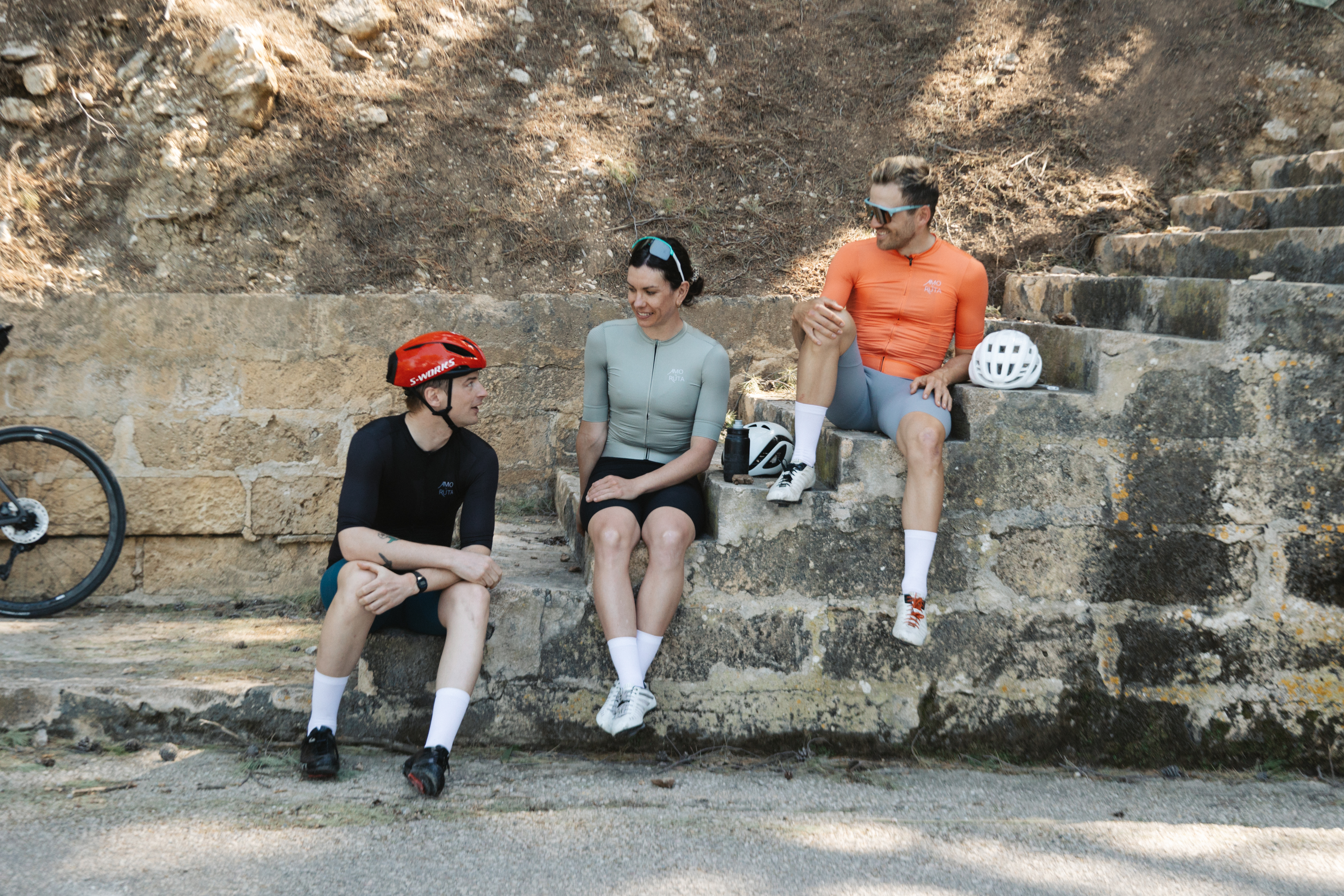 A trio of cyclists wearing Amo La Ruta gear, sitting on stone steps