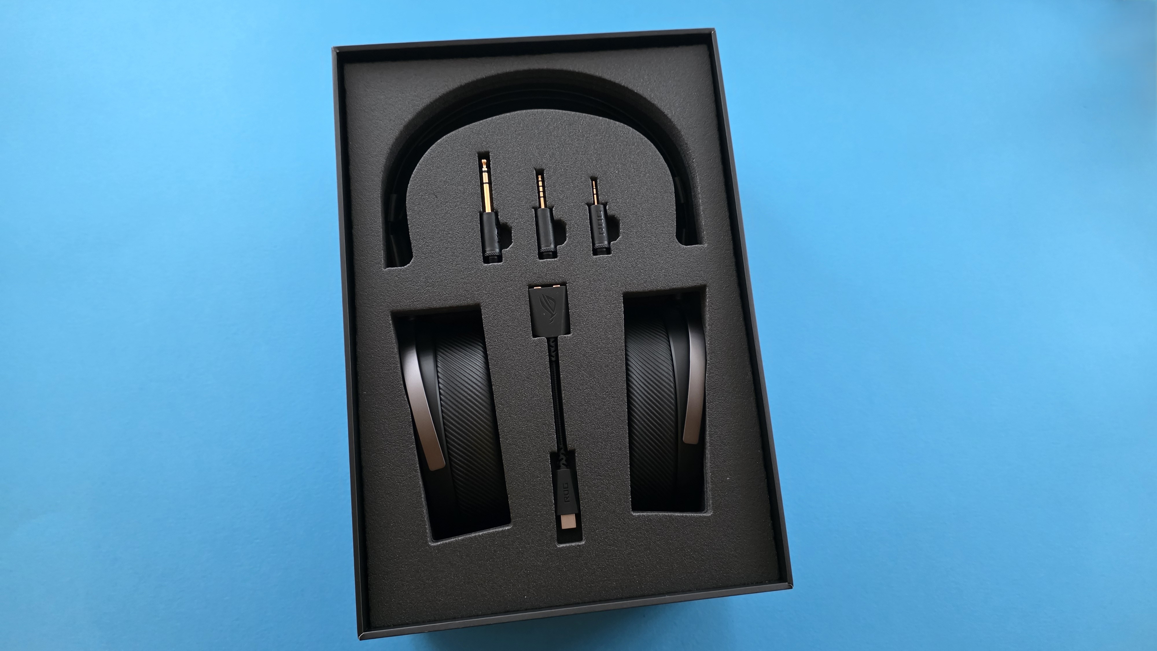 A sleek set of black headphones with surrounding foam packaging. The box includes cables and adapters, all against a bright blue background.
