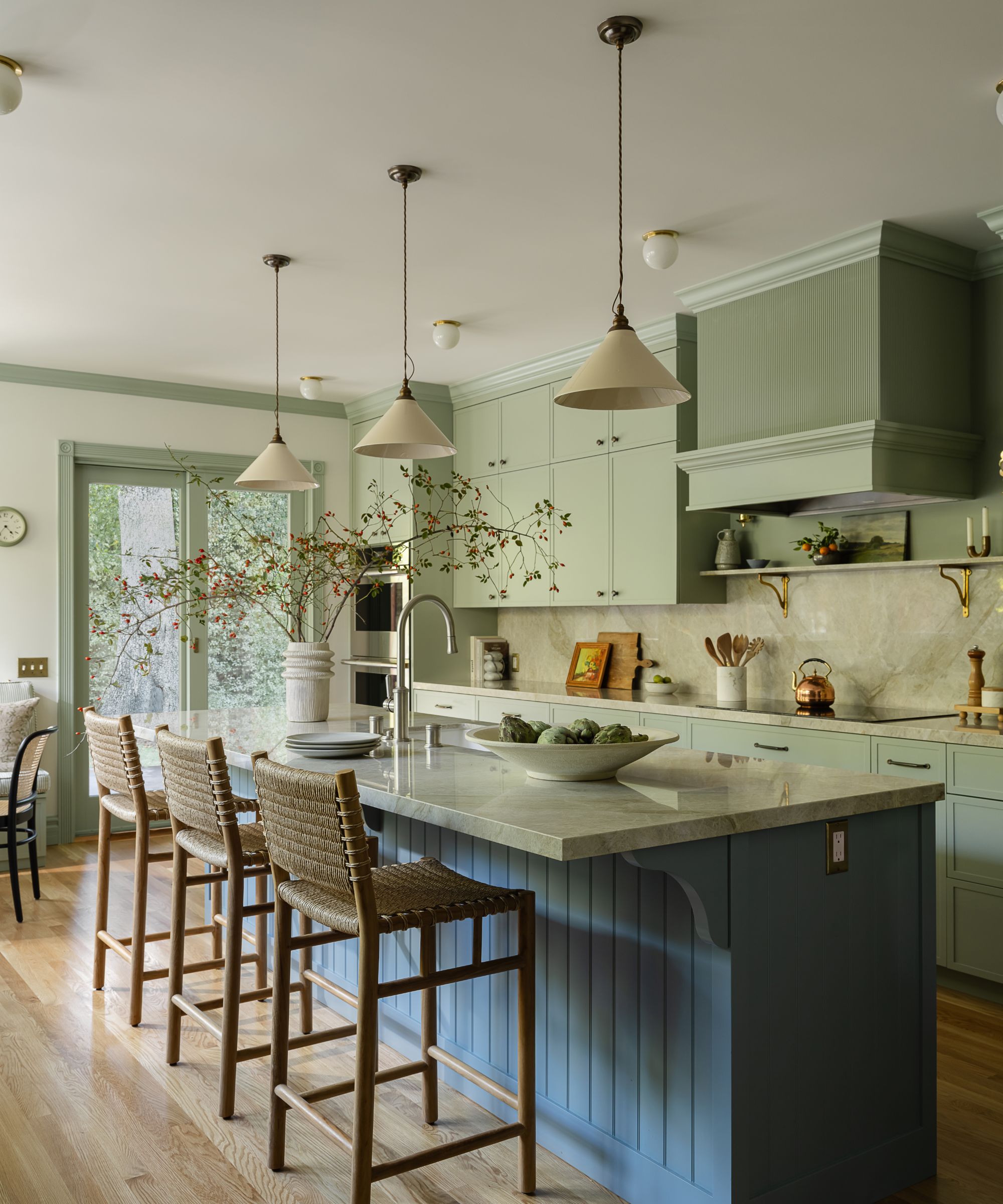 A wide view of a modern traditional kitchen featuring a large light blue island with woven barstools. The back wall is lined with sage green cabinets, a matching range hood, and a marble backsplash