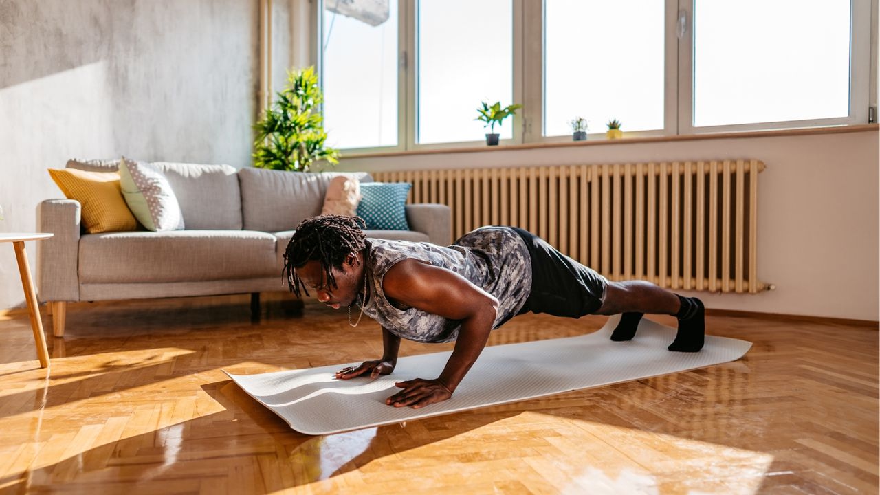 man performing a push up on an exercise mat in a living room setting with windows behind and light over him. 