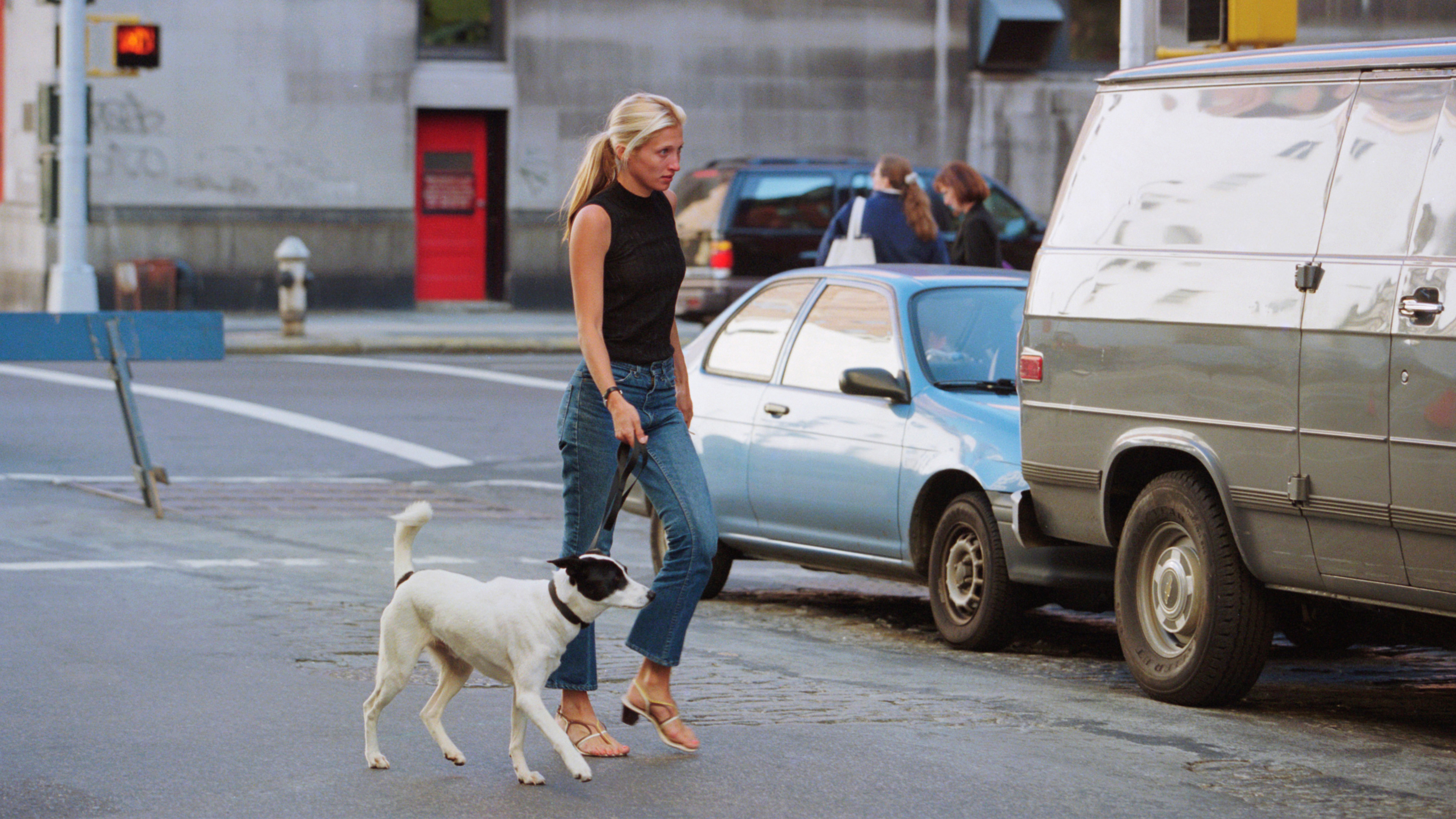 Carolyn Bessette-Kennedy wearing a black top with jeans and sandals while walking a dog.