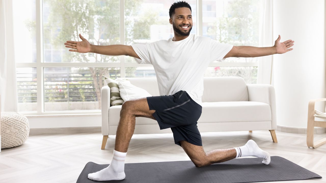 Man in living room in a lunge position with his torso rotated to one side and his arms out wide. He is smiling. He wears a white T-shirt, blue sports shorts and white socks 