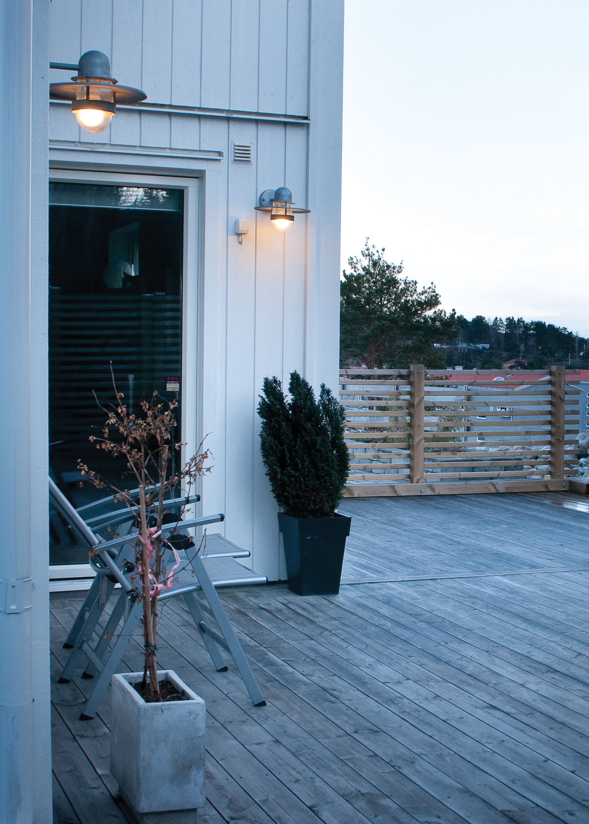 The exterior of a house at dusk with decking, a fence, and a porch light on the wall