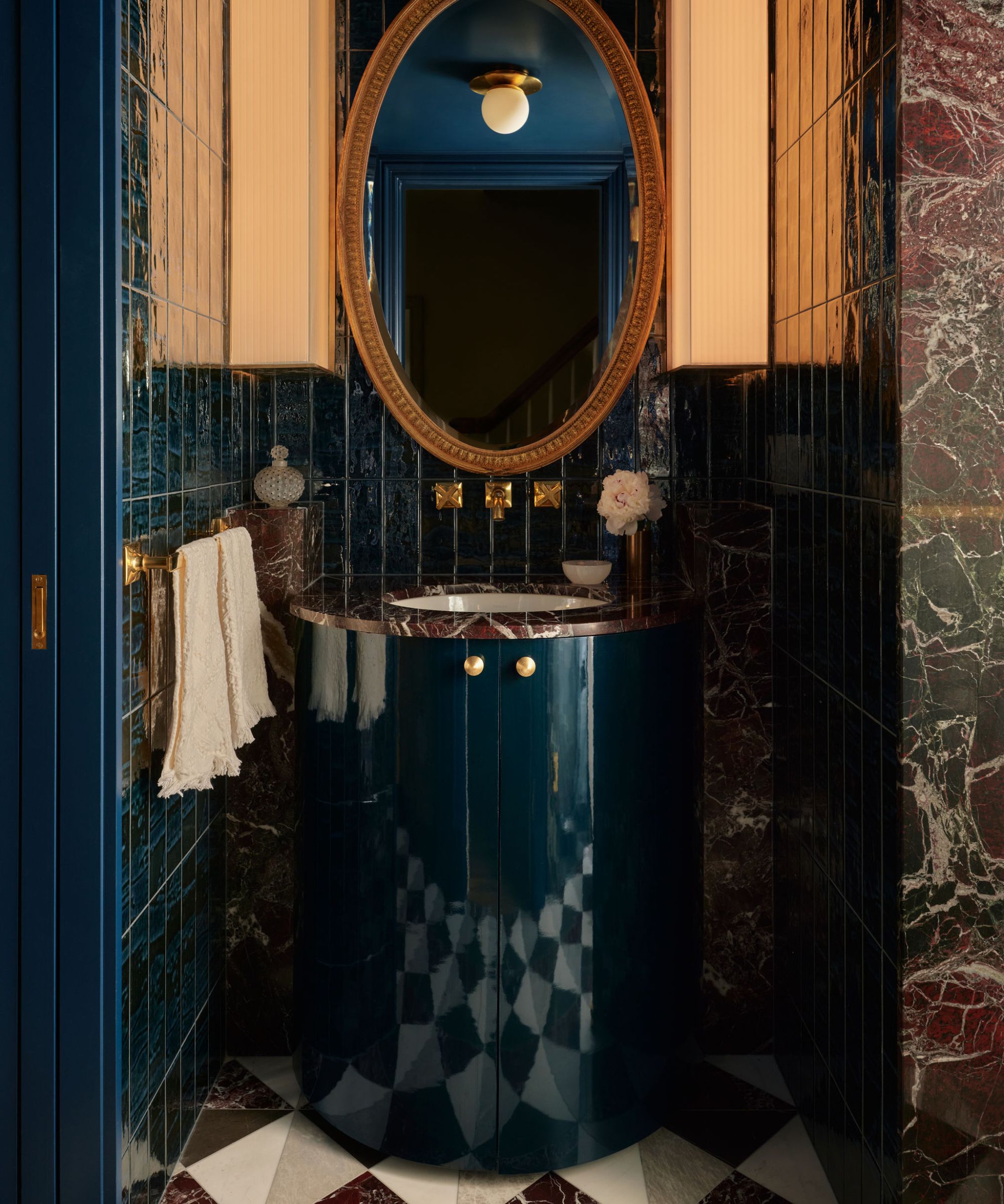 a small powder room with dark navy paint, a rounded navy vanity, rosso red marble and a large brass mirror
