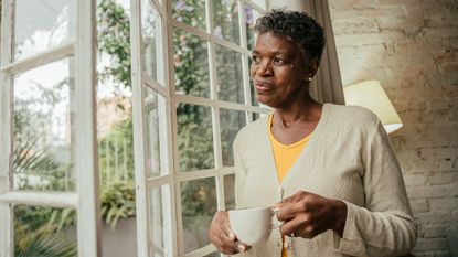 An older woman holds a coffee cup while she thoughtfully looks out her window.