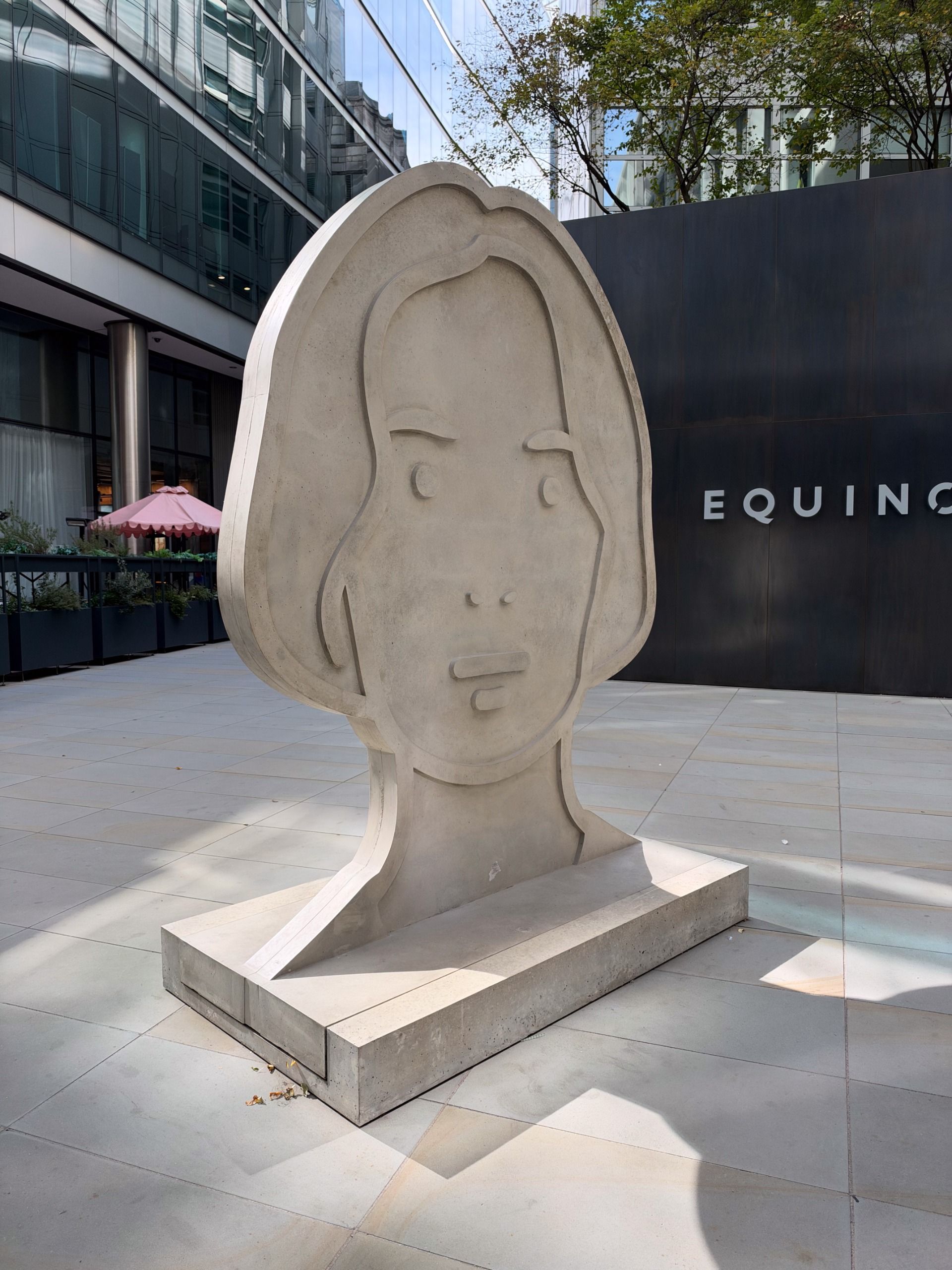 Large pale concrete sculpture of a stylized human face standing in a modern city courtyard.