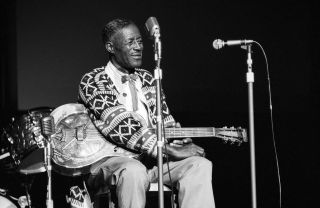 Son House sitting on a chair onstage, holding a guitar