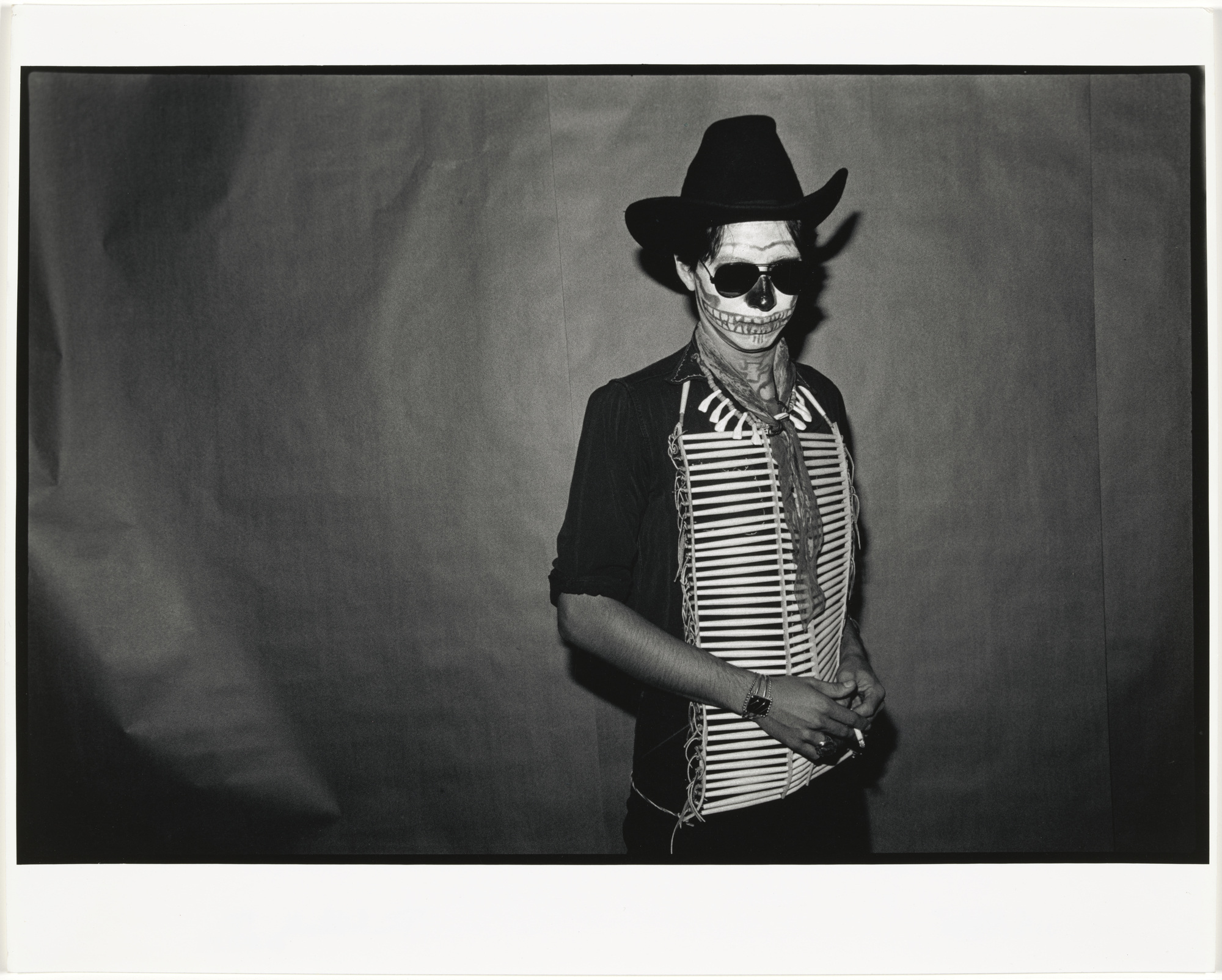 Black and white portrait of a man in a cowboy hat and sunglasses with a skull painted across his face, wearing a fringed vest and holding a cigarette.