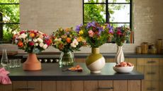 A kitchen with glossy beige tiles on the walls, large windows with black frames, black stone countertops, and a kitchen island with four colorful floral arrangements in terracotta, glass, dual-toned, and ribbed ceramic vases beside a bowl of peaches