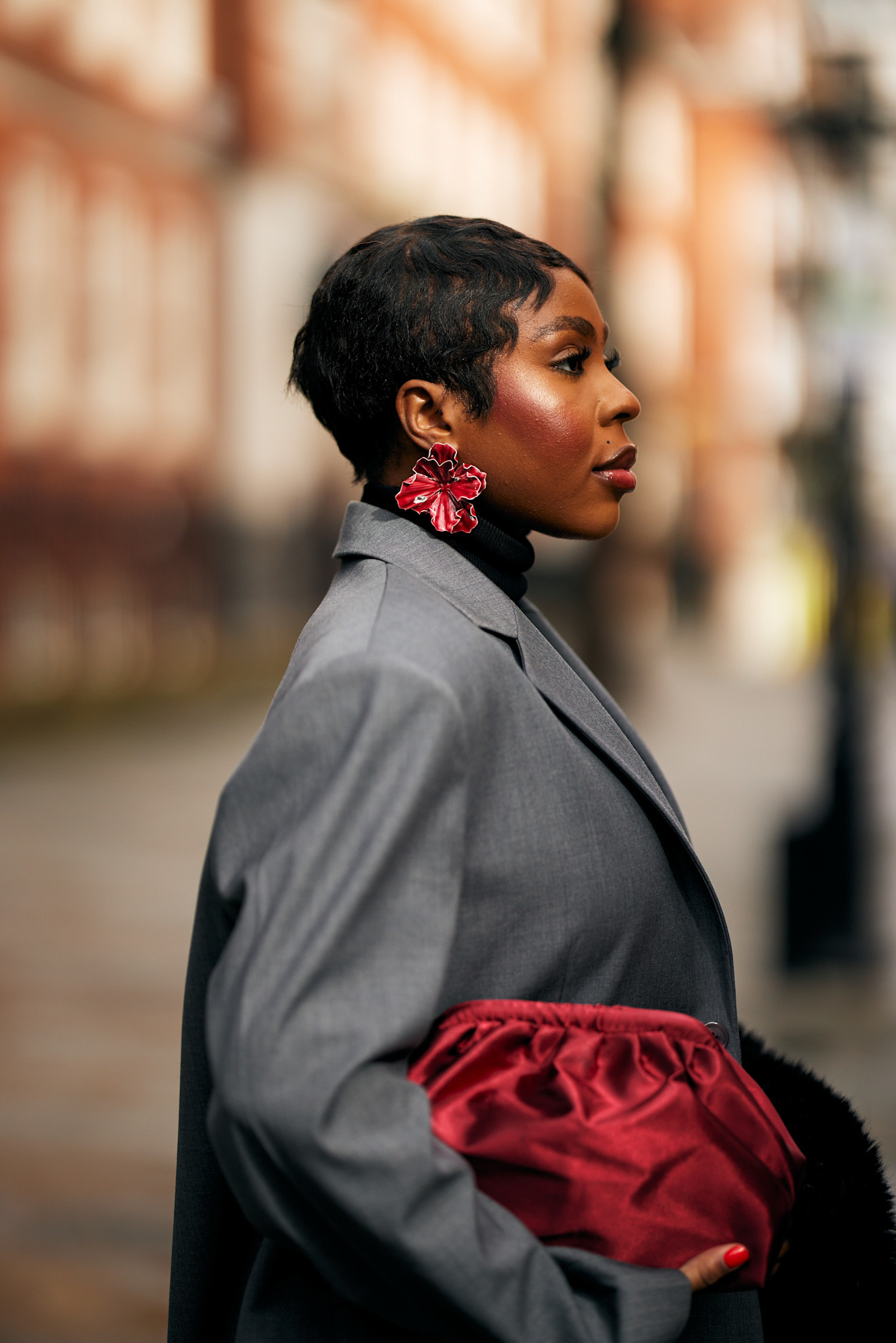 a side profile shot of a woman with dark brown hair in a pixie cut wearing a gray jacket with red flower earrings and carrying a red bag