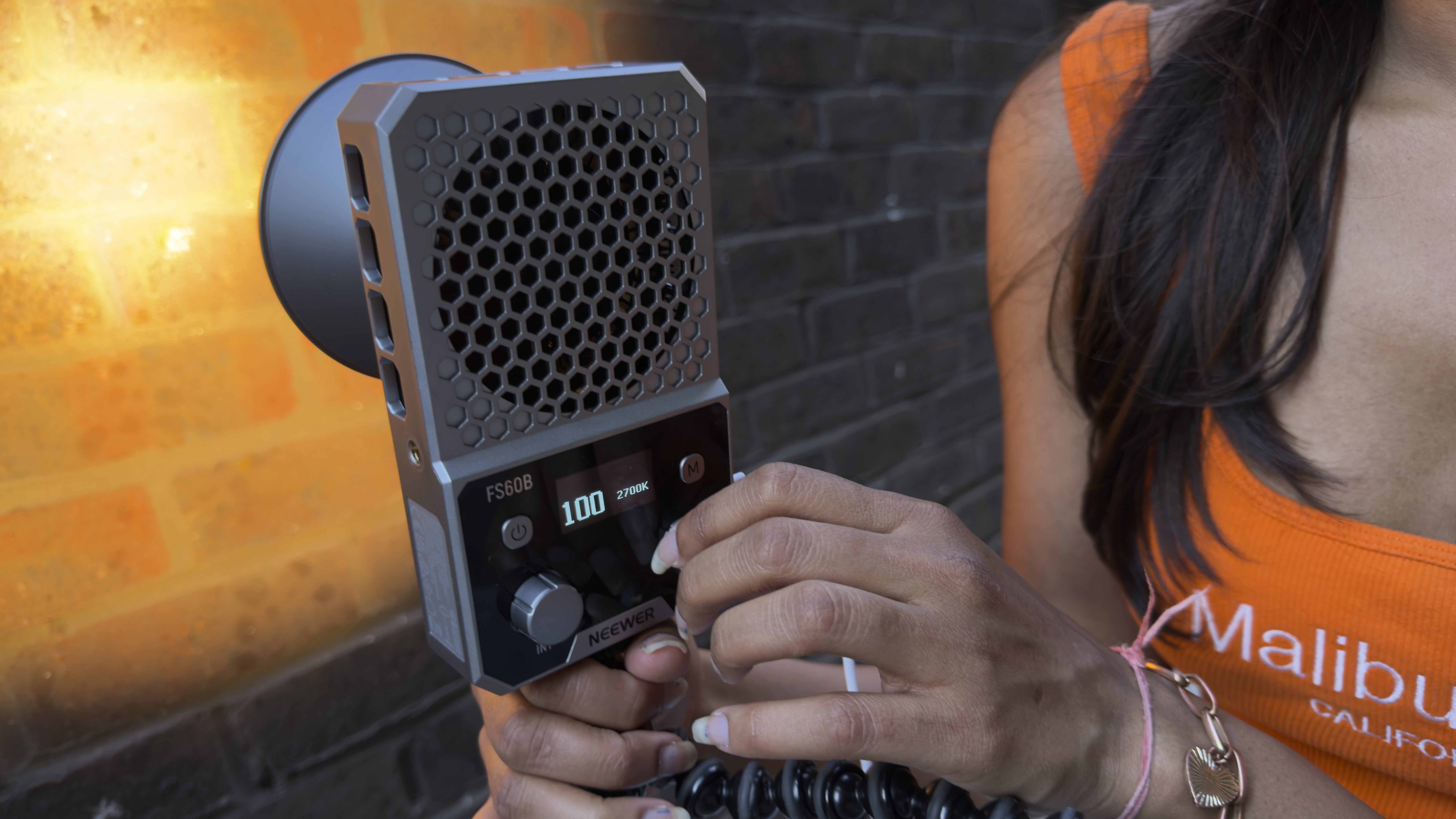Rear panel of the gray pocket-sized FS60B. A hand is twiddles a knob on the right to adjust color temperature. A brightness display shows a value of 100. The color temperature shows a value of 2700K.