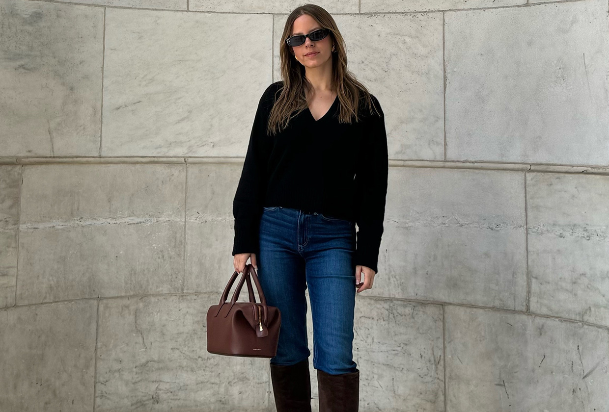 Nikki wears a black sweater, blue jeans, brown knee-high boots, and a brown bag.