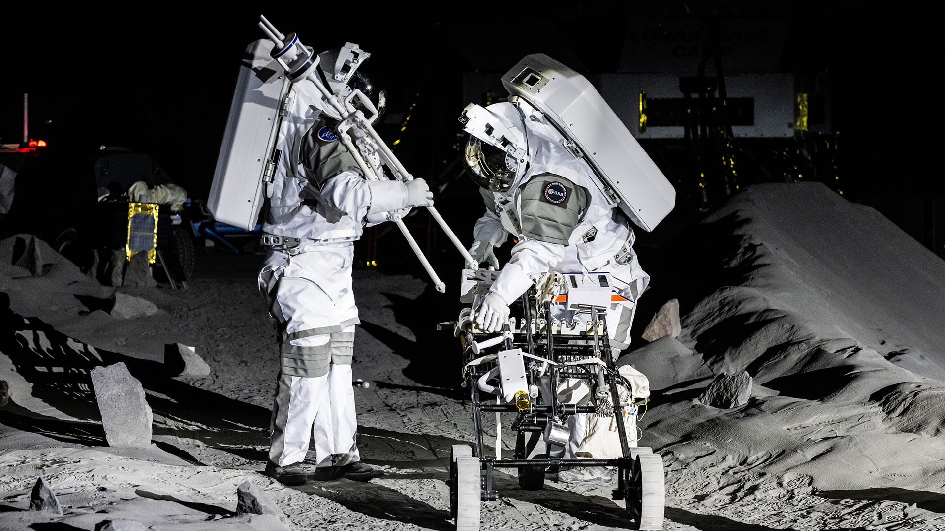 two astronauts in white spacesuits practice procedures in a simulated moon setting