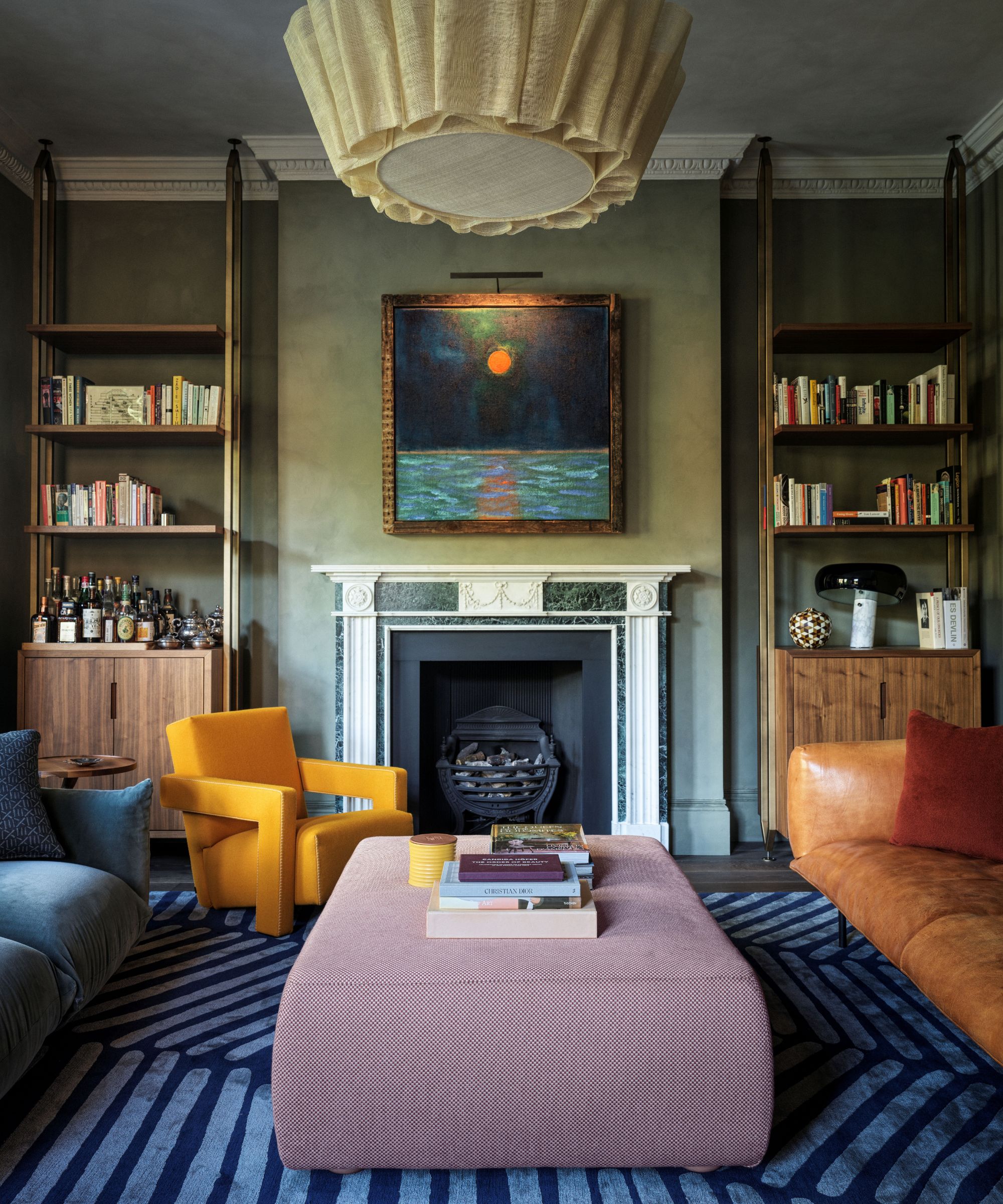 Living room with green walls, blue rug, pink ottoman, leather sofa and blue velvet sofa, yellow armchair and large fireplace with abstract painting and bookshelves on either side