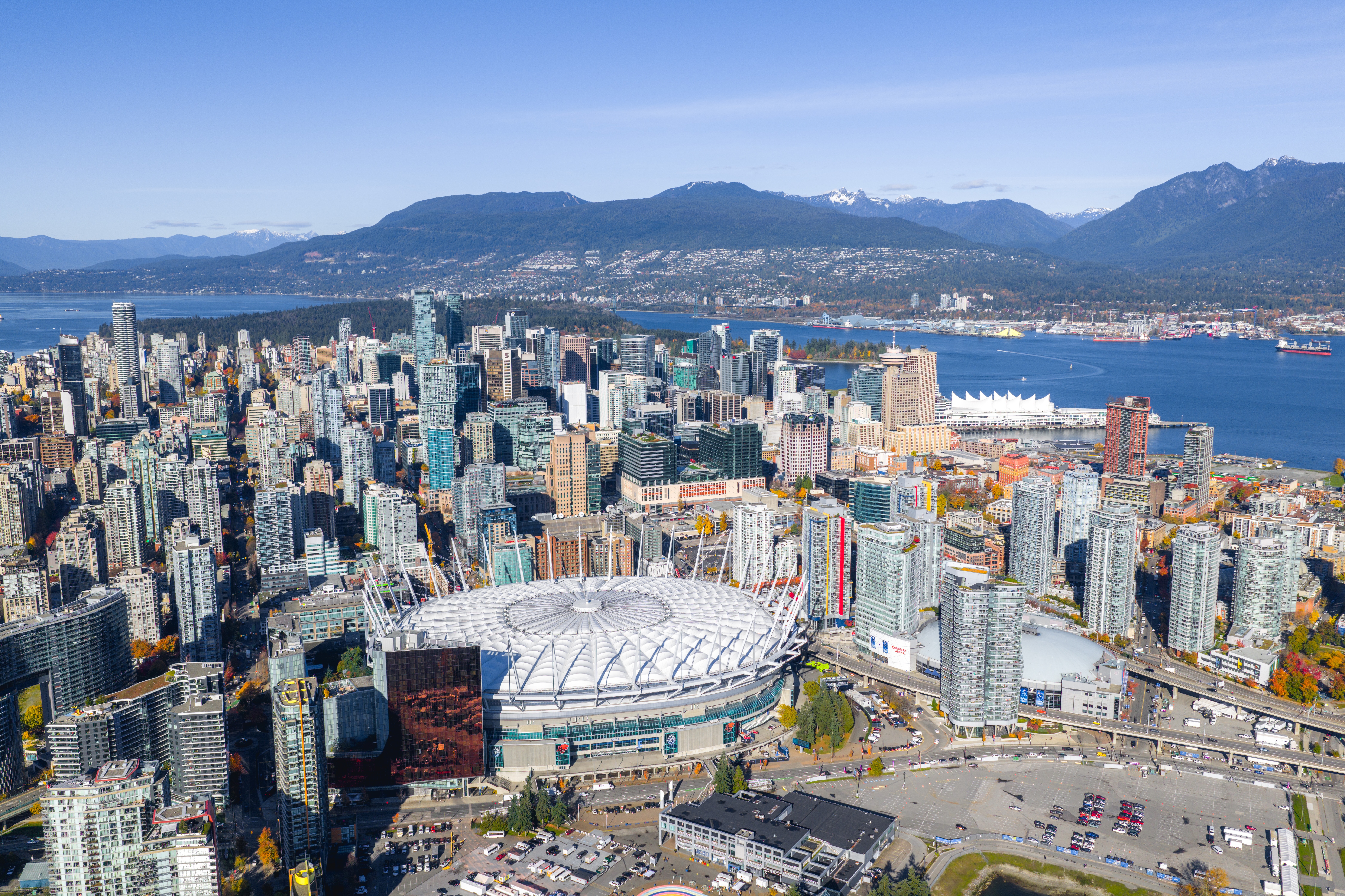 An aerial shot of downtown Vancouver in the sun, with the mountains in the distance and the city's stadium BC Place prominent in the middle of the shot