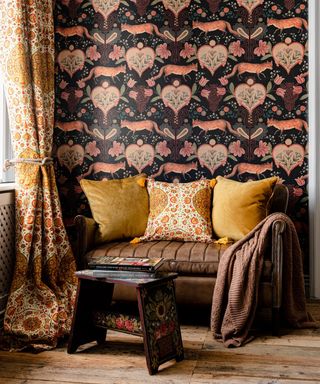 Maximalist living room design, patterned william morris style wallpaper, orange and yellow patterned curtains, small snug brown leather sofa with yellow cushions and brown throw, wooden side table with painted floral detail