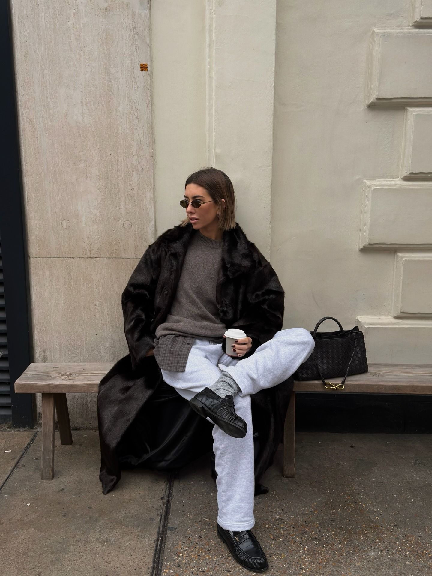 Hannah wears a fur coat, crewneck sweater, flannel shirt, gray sweatpants, and loafers.
