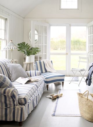 Bright white sun room with wooden patio doors and couches with blue stripes. There are wicker baskets, linen throws and a mix of metal and whitewashed furniture