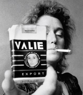 Black and white photo of a woman with a cigarette in her mouth holding a ‘VALIE EXPORT’ cigarette pack