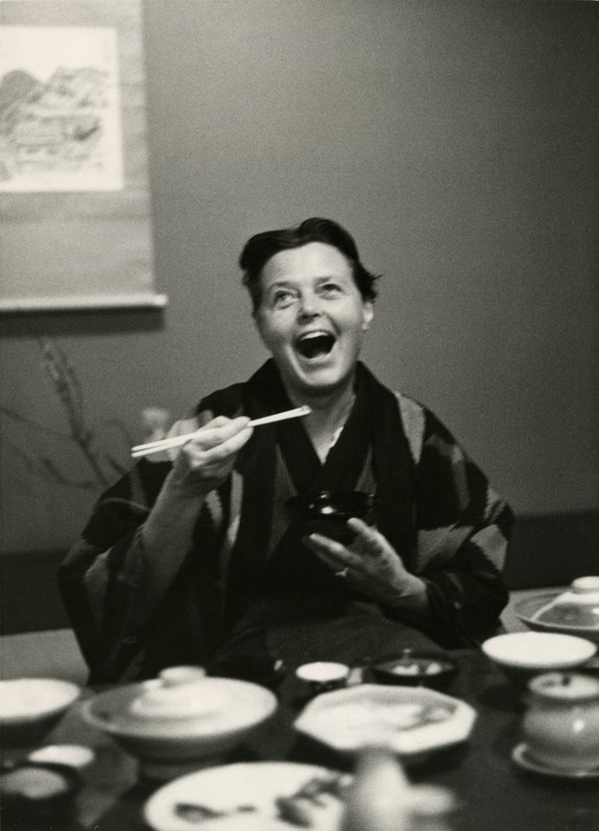 A black and white picture of designer Charlotte Perriand, dressed in traditional Japanese clothing and eating from a small bowl with a pair of chopsticks. She looks happy