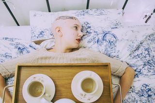 Nikki Black bald, lying in bed with tray holding 2 teacups and saucers on her chest