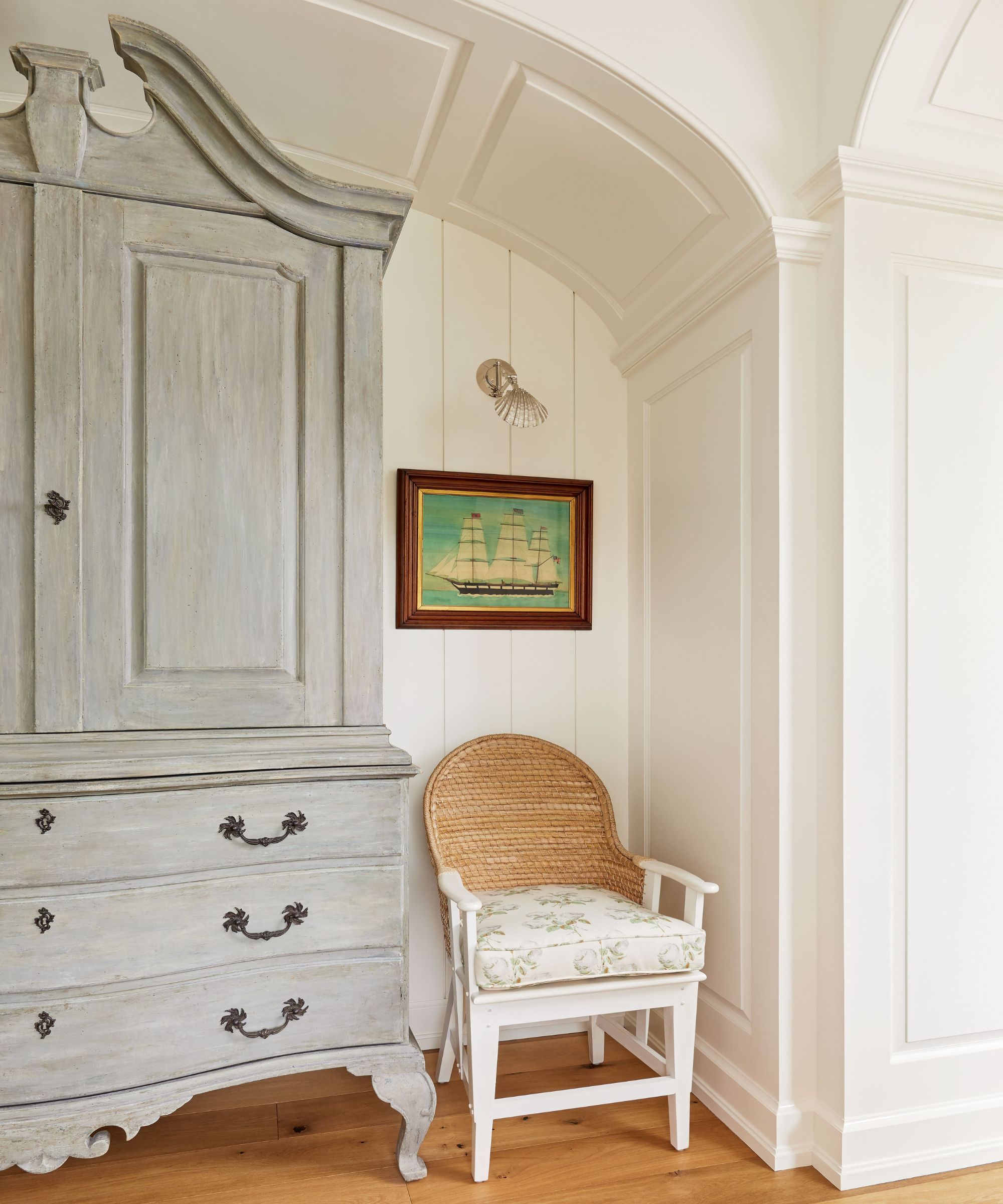 Hallway with white shiplap walls, a vintage grey armoire, a rattan white chair with a floral cushion on top, and a vintage painting of a boat with a silver shell sconce above it.