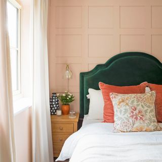 a bedroom with a peach painted panelled wall behind a dark green velvet headboards and white linen