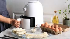 A woman's hand opening a white air fryer on a kitchen table with spring rolls and a carton of eggs. 