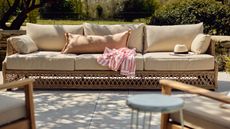 A paved concrete patio with a rattan three-seater outdoor sofa with cream cushions.