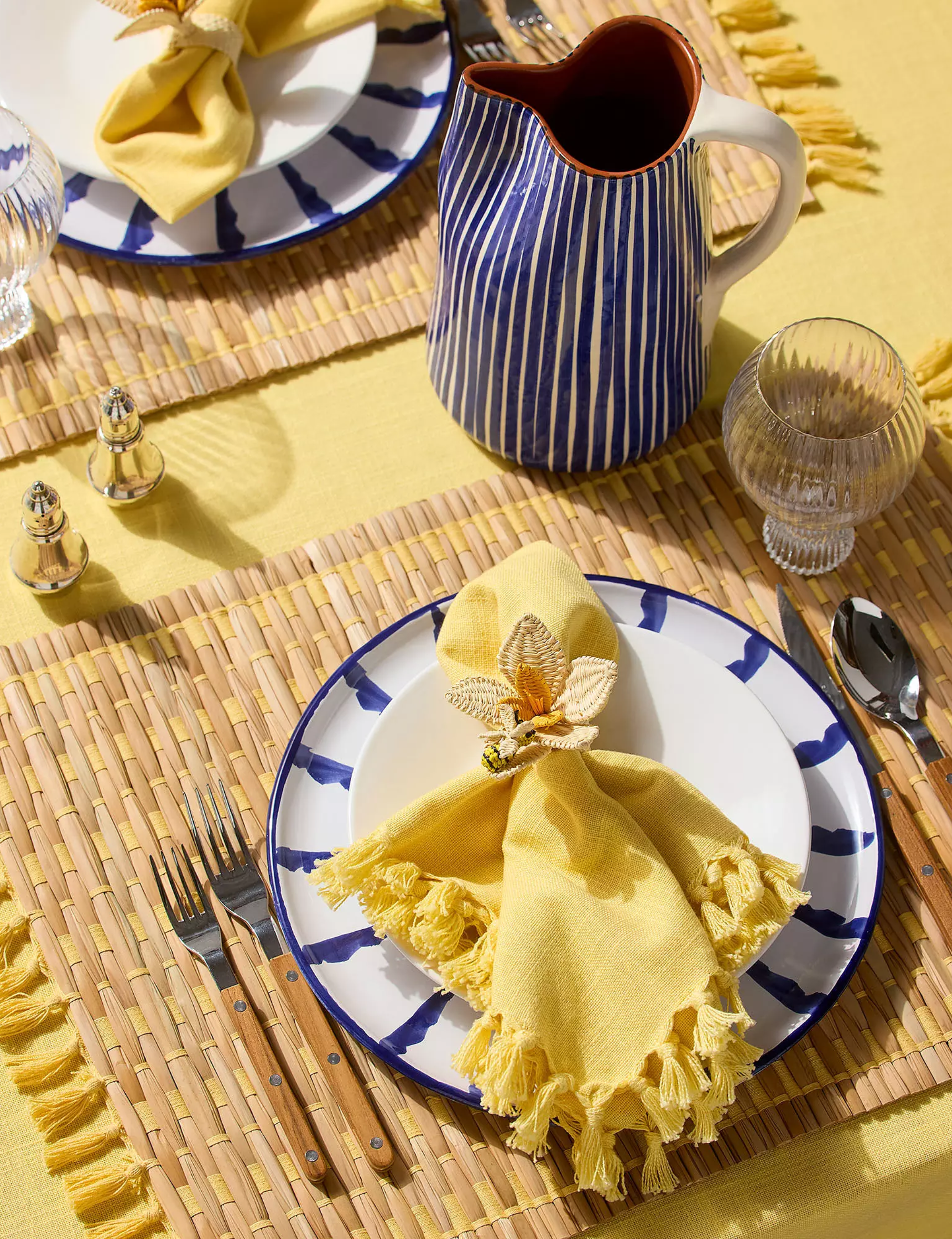 Cheerful yellow-and-navy outdoor tablescape featuring tassel-bordered napkins, woven napkin rings, and striped ceramics