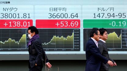 Pedestrians walk past a board with Japanese stocks