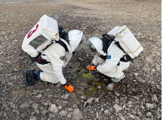 two people in space suits pick up rocks outside