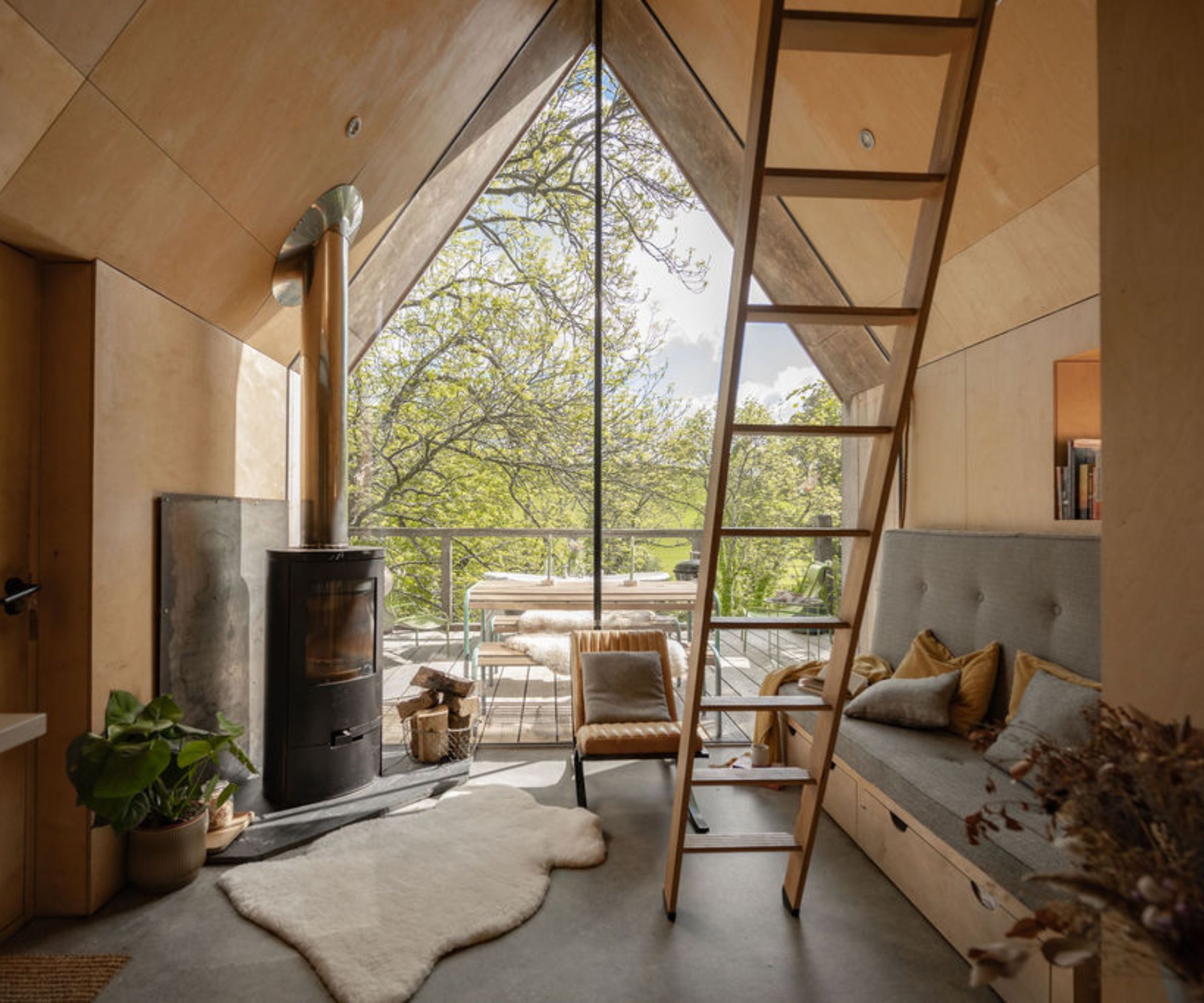 Scandinavian-style treehouse interior with a soaring pitched ceiling and minimalist plywood walls. A ladder leads to a loft bed, while a log-burning stove and floor-to-ceiling windows connect the cosy living space to the surrounding treetops.