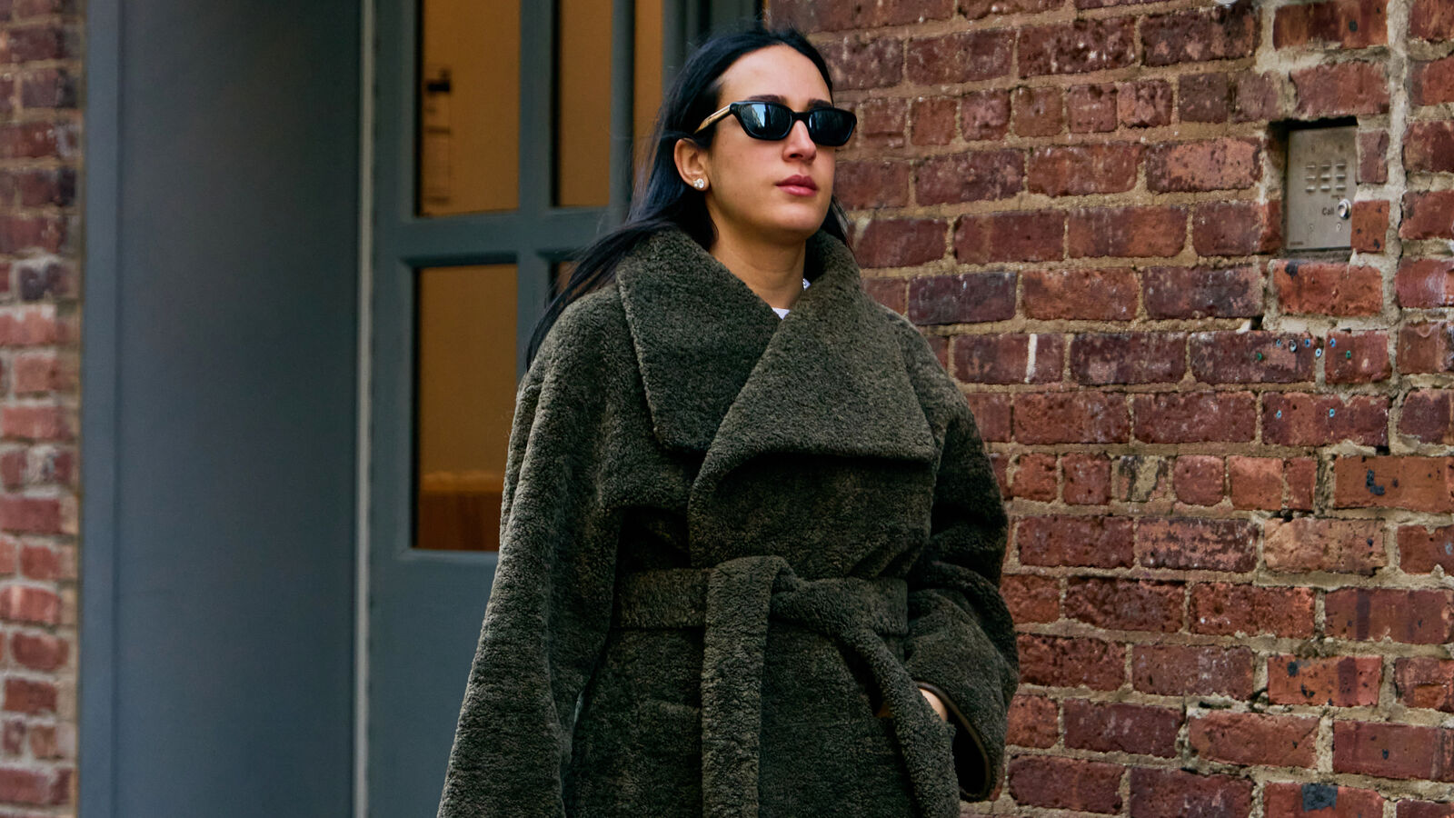 Woman wears green faux fur coat while carrying a black quilted handbag and tan sneakers. 