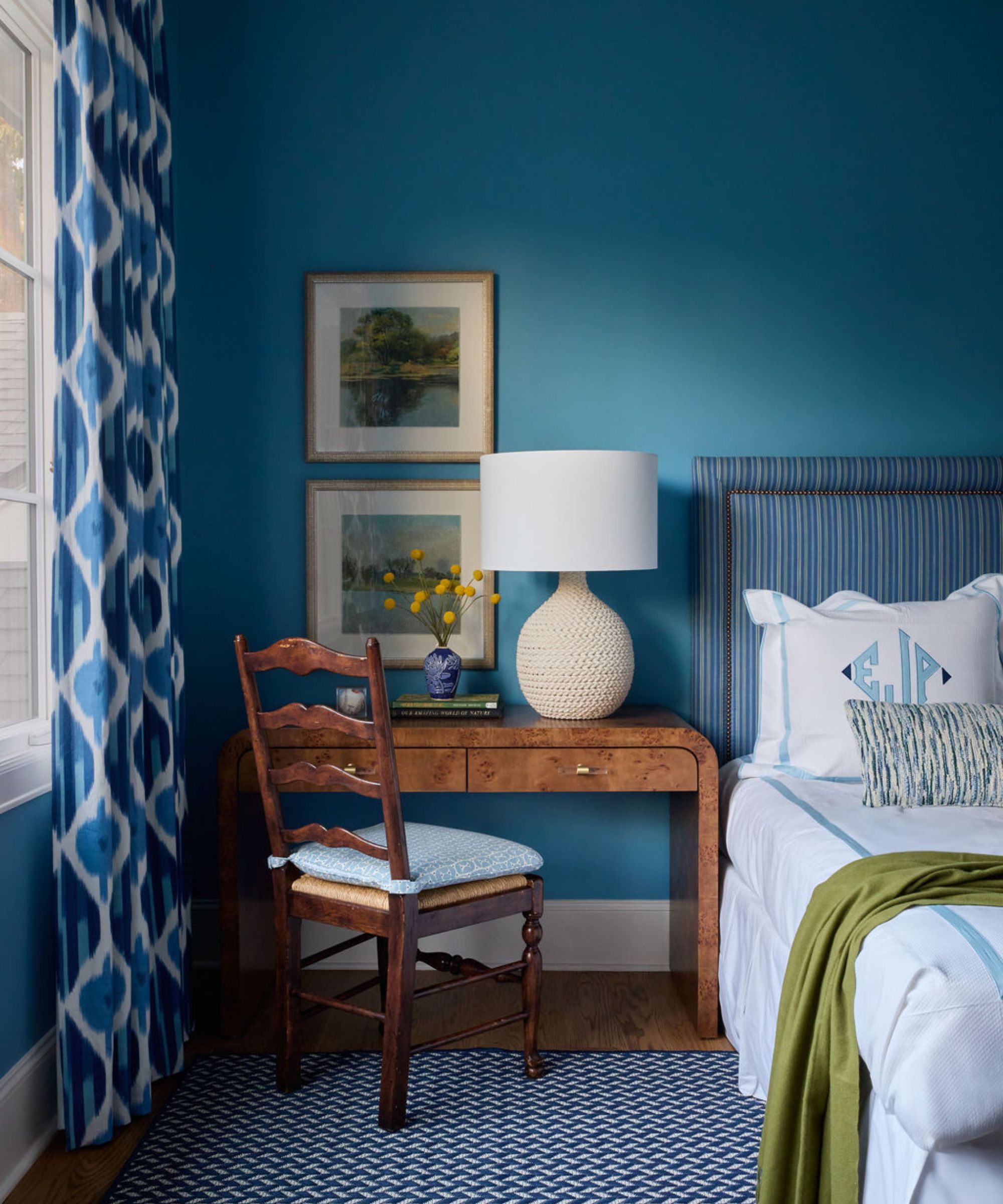 A corner of a cozy bedroom with mid-tone blue walls, a navy striped headboard with crisp white bedding and a lime green throw, a burl wood table with a wooden chair, a lamp, and two framed artworks. A blue geometric-patterned rug and blue patterned curtains.