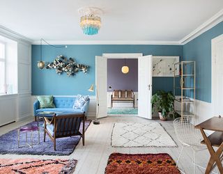 View of a 2013 living area at The Apartment gallery featuring wood flooring, blue walls with white wood panelling at the bottom, a blue sofa with cushions, a round purple and metal side table, a wooden desk, chairs, rugs in different colours, a pendant light, a chandelier, a window, a green plant in a pot, a shelving unit and wall art. Another space with purple walls, a pendant light, a chair and a green rug can be seen through open white doors