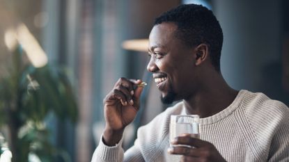 Man is about to put a vitamin pill in his mouth as he holds a glass of water in his other hand