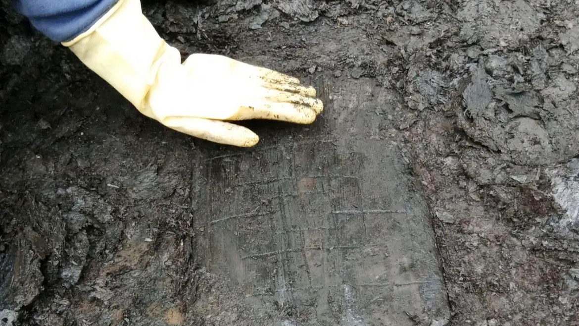 a person with a yellow-gloved hand touches a waterlogged rectangle of wood that has three concentric squares carved into it