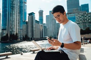 A person using a smartphone and laptop outdoors with modern buildings and water in the background.