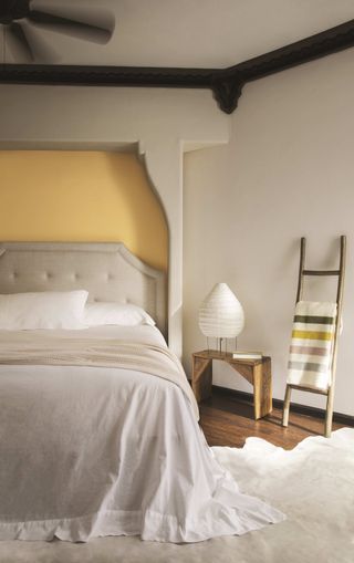 bedroom with taupe and yellows walls, upholstered bed, off white bedlinen, stool, ladder, wooden floor, rug