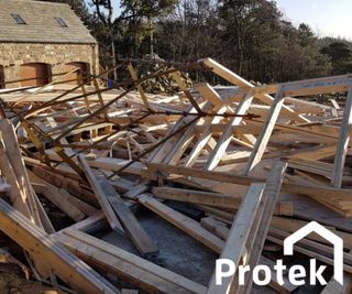 A storm-damaged site, with a timber frame for a new home strewn across the site. there is a woodland and a garage in the background.